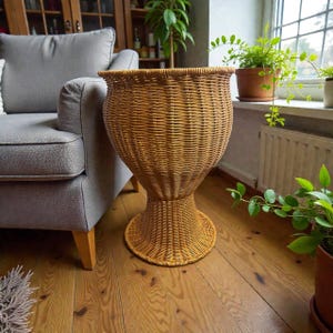 Puede incluir: Un soporte para plantas alto con forma de reloj de arena, fabricado en mimbre tejido de color marrón claro. La pieza presenta una base circular ancha que se estrecha hacia una sección central antes de expandirse en un gran cesto redondeado.