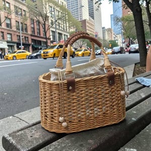 Puede incluir: Un bolso de mimbre tejido con un interior de tela clara. El bolso tiene un asa y detalles de cuero marrón, con detalles de cuentas de madera en el cierre de cordón. El bolso está sobre un banco de madera con una calle de la ciudad al fondo.