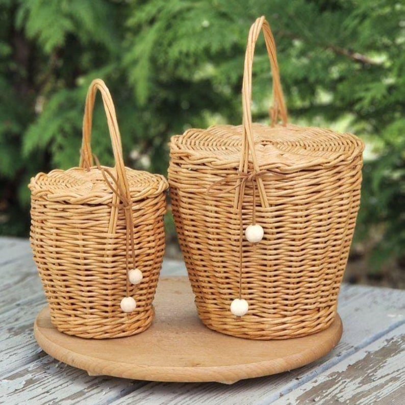 May include: Two light brown woven wicker baskets with lids and vertical handles. Each basket features a thin brown cord tied to the handle, decorated with two small, round, off-white wooden beads hanging down the side of the basket body.