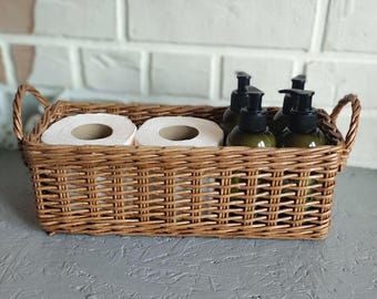 May include: A rectangular brown woven wicker basket with two side handles containing two white toilet paper rolls and three dark green plastic pump bottles with black dispensers, arranged in a row inside the storage container.