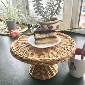 May include: A round wicker cake stand with a slice of chocolate cake on a white plate. The stand has a pedestal base. Potted plants and a pink rose are visible in the background. The cake stand is made of woven wicker.