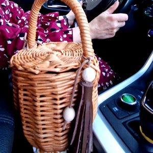 May include: A cylindrical woven wicker basket bag with a sturdy arched handle. The bag features a lid and is decorated with a brown leather tassel attached to a cord with two light-colored wooden beads hanging from the side of the handle.