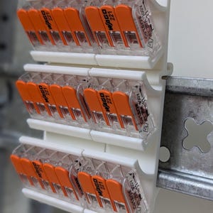 Può includere: Primo piano di tre file di connettori elettrici. Ogni connettore ha un alloggiamento in plastica trasparente con leve arancioni e la scritta "WAGO 221". I connettori sono montati su una base di plastica bianca.