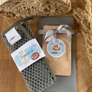 Könnte beinhalten: Ein Laib knuspriges Brot liegt auf einem Holzschneidebrett neben einem Netz-Reinigungstuch mit dem Text "Teigwisch". Ein kleines Paket mit Schleife und rundem Etikett mit dem Text "LAKROO" und "Sauerteigstarter" ist ebenfalls vorhanden.