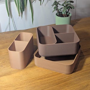 Pode incluir: Uma coleção de organizadores de mesa em tons neutros. O conjunto inclui um porta-lápis dividido, uma bandeja retangular e um organizador maior com vários compartimentos. Os organizadores são em tons de castanho e bege, com uma textura canelada em algumas peças.