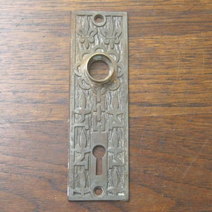 Peut inclure: Plaque de porte antique en bronze avec trou de serrure et motifs floraux et géométriques décoratifs. La plaque rectangulaire comporte une ouverture circulaire pour la poignée de porte et est montée sur une surface en bois.