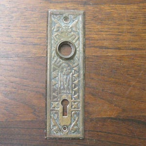 Peut inclure: Plaque de porte en laiton antique avec des détails en relief ornés. La plaque rectangulaire présente un trou de serrure, une ouverture circulaire et des motifs décoratifs. Le laiton a un aspect vieilli, suggérant l'âge et l'utilisation. La plaque est fixée sur une surface en bois.