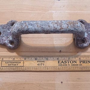 May include: A weathered, grey and brown metal door handle with a textured, rusted finish and four mounting holes. The handle is positioned above a wooden ruler, showing a total length of approximately 24.1 cm from end to end.