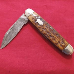 May include: An open pocket knife with a worn, silver blade and a handle featuring a brown, textured pattern. The handle has silver accents and a small, silver emblem. The knife rests on a red surface.