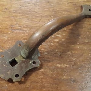 Peut inclure: Une poignée de porte en bronze antique avec une poignée incurvée en bois. La poignée a une plaque arrière décorative avec une ouverture carrée et plusieurs trous de montage. Le bois est brun chaud, le métal montre des signes d'usure.
