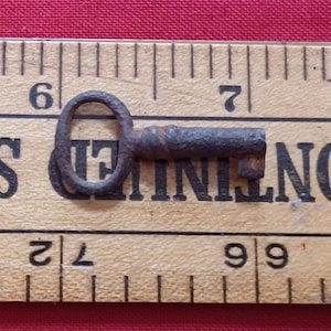 Peut inclure: Une petite clé ancienne avec une poignée circulaire et un panneton rectangulaire. La clé est en métal sombre et rouillé, posée sur une règle en bois. La règle a des marques et des chiffres noirs.
