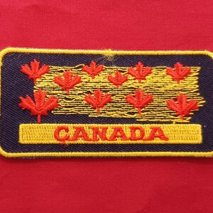 May include: A rectangular patch with a navy blue border and a yellow embroidered outline. The patch features red maple leaves and the word "CANADA" in yellow lettering. The background is red.