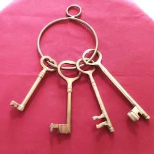 Peut inclure: Un ensemble de quatre clés anciennes en laiton, attachées à un grand anneau métallique. Les clés présentent des formes et des tailles différentes, avec des détails complexes. Elles sont disposées sur un fond rouge.