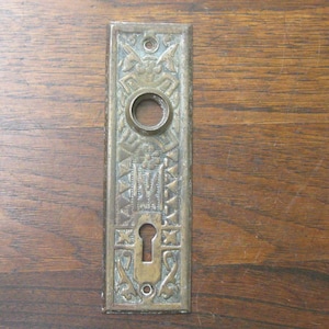 May include: An antique brass door plate with detailed embossed patterns. The rectangular plate includes a keyhole, a circular opening, and decorative designs. The aged metal has a weathered look, indicating vintage craftsmanship.