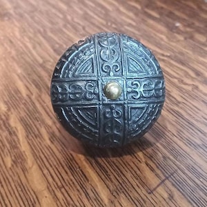 Peut inclure: Un bouton de meuble rond en métal sombre avec un motif en relief orné. Le bouton présente un goujon central de couleur or et un motif détaillé de lignes entrecroisées et d'éléments décoratifs. Le bouton est posé sur une surface en bois.