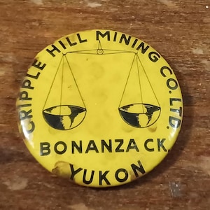 May include: A circular yellow button featuring black text and a scale design. The text reads "CRIPPLE HILL MINING CO. LTD." and "BONANZA CK YUKON". The button has a vintage, worn appearance.