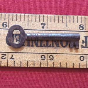Peut inclure: Une petite clé ancienne en métal, avec une tête ovale et une tige cannelée. La clé mesure environ 6,35 cm de long et repose sur une règle en bois avec des marques noires. Le fond est de couleur rouge uni.