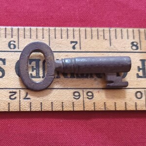 May include: A small, antique key with an oval head and a cylindrical body. The key is made of a dark, possibly rusted metal and rests on a wooden ruler. The ruler has black markings and numbers. The background is a solid red fabric.