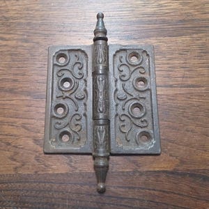 Peut inclure: Une ancienne charnière en fonte avec des volutes décoratives et une goupille centrale. La charnière est de couleur métal foncé et vieilli, avec des signes d'usure. La charnière mesure environ 10 cm de haut et 7,6 cm de large.
