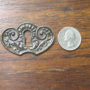 May include: An antique brass keyhole cover with an ornate, heart-shaped design. The cover features a central keyhole and is next to a U.S. quarter coin for size comparison. The keyhole cover is likely from a vintage cabinet or furniture piece.