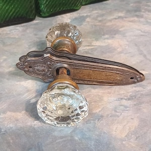 May include: A vintage door handle set featuring two clear, faceted glass knobs attached to an elongated, ornate brass backplate with a weathered patina. The metal plate displays intricate embossed detailing and screw holes at both ends.