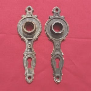 Peut inclure: Deux plaques de porte anciennes en bronze. Chaque plaque a un design orné avec un trou de serrure et une ouverture circulaire. Les plaques sont orientées verticalement et ont une finition vieillie.