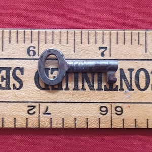 Peut inclure: Une petite clé en métal antique avec une tête ovale et une tige cannelée. La clé mesure environ 5 cm de long et repose sur une règle en bois avec des marques noires. Le fond est un tissu rouge uni.