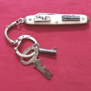 Peut inclure: Un porte-clés vintage avec un couteau de poche, doté d'un manche crème et d'une lame en métal argenté. Le couteau présente un petit relief du Mont Rushmore et l'inscription "Mt. Rushmore". Le porte-clés comprend une clé et un porte-clés.