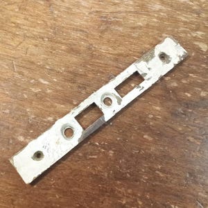 May include: A rectangular, metal door latch plate with a distressed, off-white painted finish. The plate has two rectangular cutouts and three circular holes for screws. The paint is chipped, revealing the metal underneath.