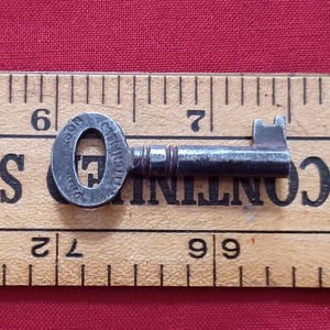 Peut inclure: Une clé en métal gris foncé avec une tête circulaire et un panneton rectangulaire. La clé est posée sur une règle en bois avec des marquages noirs. La règle est placée sur une surface rouge. La tête de la clé porte l'inscription "C.P. 1912".