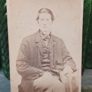 May include: A sepia-toned portrait of a man in a suit, seated and holding a hat. The man is wearing a dark jacket over a vest and a patterned necktie. The photograph has a vintage aesthetic.