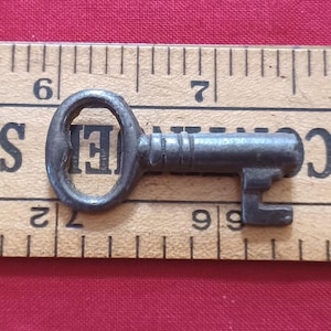 May include: A small, antique metal key with an oval head and a rectangular bit. The key is dark grey and appears aged. It rests on a wooden ruler with black markings and numbers. The ruler is on a red surface.