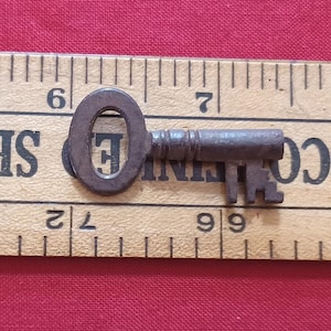 Peut inclure: Une petite clé ancienne en métal sombre et vieilli. La clé a une tête ovale et un panneton détaillé. Elle repose sur une règle en bois avec des marquages noirs, sur fond rouge.