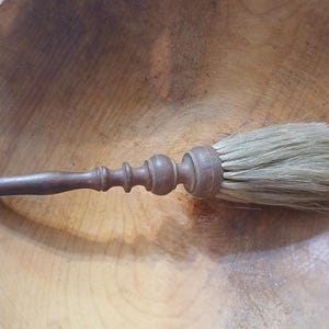 May include: A wooden pastry brush with a turned handle and a small metal loop at the end. The brush features light-coloured natural bristles securely attached to the dark brown, carved wooden handle, which has a series of decorative rounded ridges.