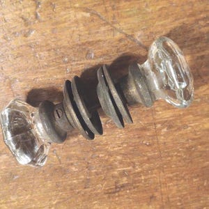 May include: Vintage door knob made of clear glass and metal. The knob has two clear, faceted glass ends, connected by dark metal washers and a central metal rod. The hardware is designed for a door.