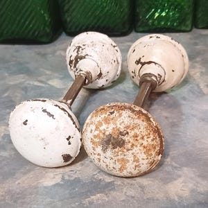 May include: Two antique door knobs with white and brown distressed finishes. The knobs have a round shape and are attached to a metal rod. The knobs are likely made of ceramic or porcelain.