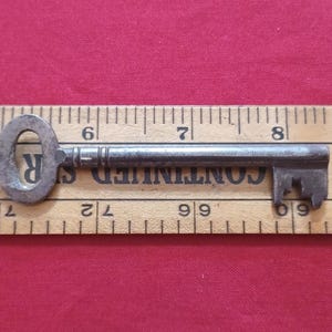 May include: An antique metal key with an oval head and a rectangular bit. The key is approximately 7.6 cm long and lies on a wooden ruler with black markings. The key is a dark, aged metal colour.