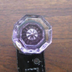 May include: A close-up of a vintage glass doorknob with an octagonal shape. The knob is a light purple colour and has a detailed, starburst-like design in the centre. The metal base is black with a screw visible.