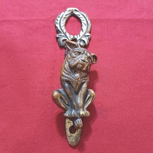 May include: A vertical brass door knocker featuring a sculpted dog figure in a seated position. The top of the piece has a circular ring with a leaf-like pattern, while the bottom section includes a small circular hole for mounting hardware.
