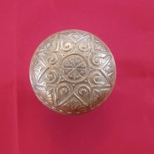 Peut inclure: Un objet rond argenté avec un motif complexe de volutes, d'étoiles et un motif circulaire central. L'objet est posé sur un fond rouge, mettant en valeur ses détails ornés.