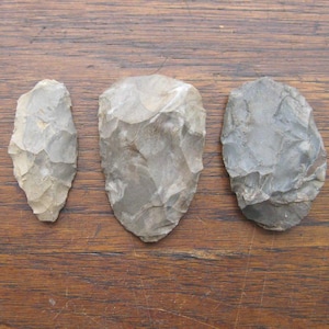 May include: Three grey, hand-knapped stone tools, possibly arrowheads or blades, are displayed on a wooden surface. The tools vary in size and shape, with a rough, textured appearance. The background is a warm-toned wooden surface.