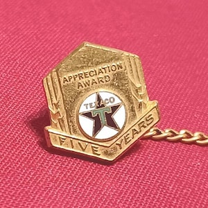 May include: A gold-toned metal lapel pin shaped like a shield, featuring the text APPRECIATION AWARD and FIVE YEARS. In the centre is a circular emblem with a black star, the word TEXACO, and a green letter T, attached to a gold chain.