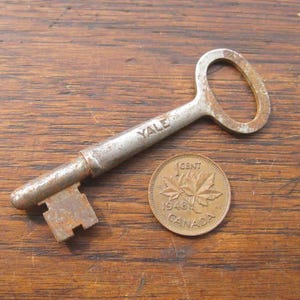 May include: A vintage Yale key and a Canadian one-cent coin from 1948 on a wooden surface. The key shows wear and tear, and the coin has a maple leaf design. The key is approximately 7.6 cm long.