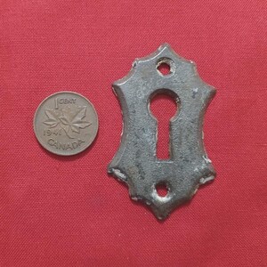 May include: A dark grey, ornate metal keyhole cover with a keyhole shape and decorative edges. A Canadian one-cent coin from 1941 is next to the keyhole cover. The keyhole cover is on a red surface.