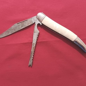 Peut inclure: Un couteau de poche pliant avec un manche lisse blanc cassé et deux lames en métal. Une lame est un couteau pointu standard, tandis que l'autre est un outil dentelé avec une base crantée. Les deux lames présentent des signes d'oxydation.