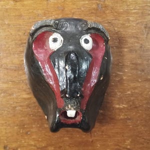 May include: A black mask with red detailing around the eyes and mouth, featuring white eyes and a single tooth. The mask appears to be carved, possibly from wood, and has a worn, distressed finish, giving it a vintage or rustic appearance.
