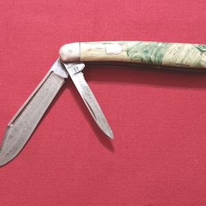 Peut inclure: Un couteau de poche pliant avec deux lames en métal déployées. Le manche présente un motif marbré vert et crème avec des mitres métalliques argentées aux deux extrémités. Une petite incrustation en forme de bouclier est visible au centre.