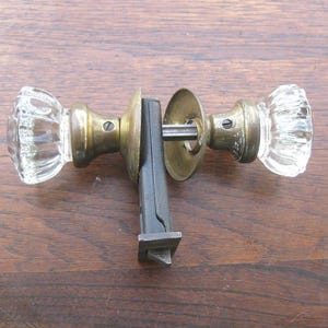 May include: A vintage door knob set featuring clear glass knobs with a ribbed design and brass fittings. The set includes the spindle and a black metal latch mechanism. The door knob set is displayed on a wooden surface.