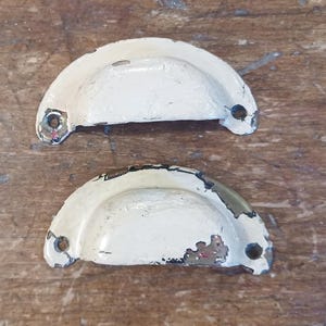May include: A pair of vintage white metal drawer pulls with a distressed finish. Each pull is a half-circle shape with two holes for mounting. The paint is chipped, revealing the metal, giving them a rustic appearance.