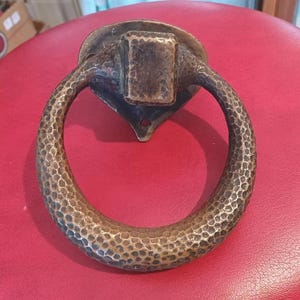May include: A bronze-toned metal door knocker featuring a circular ring with a hammered, textured surface. The knocker includes a central square mounting block and a heart-shaped backplate with visible screw holes for attachment to a door surface.